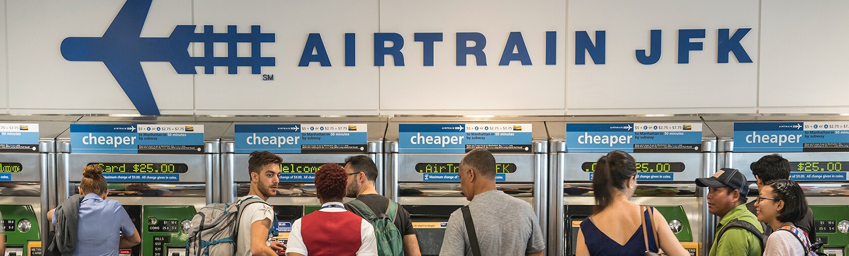 A sign saying AIRTRAIN JFK stretches across a wall above a line of MetroCard vending machines. People with backpacks and luggage are gathered near the ticket machines.