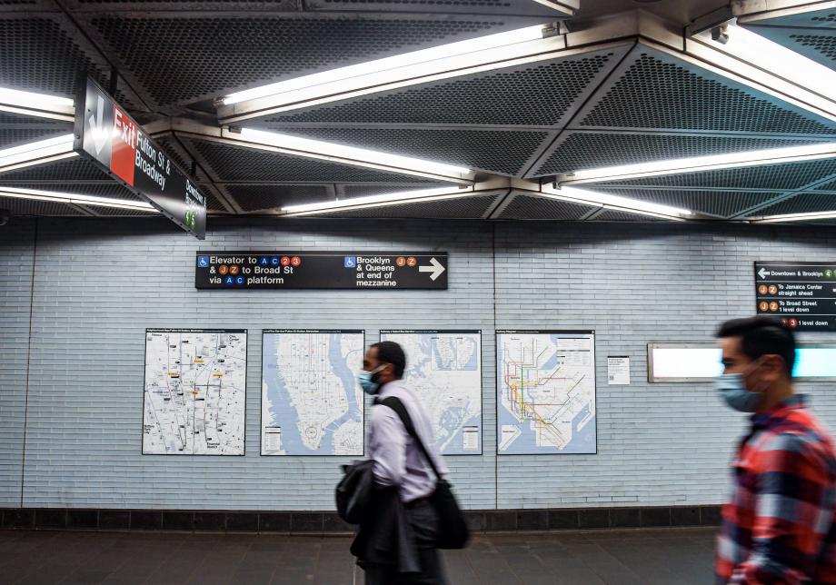 Four transit maps are installed on the wall of a subway station. Two men are walking by, one in a white shirt and the other in a red checked shirt.