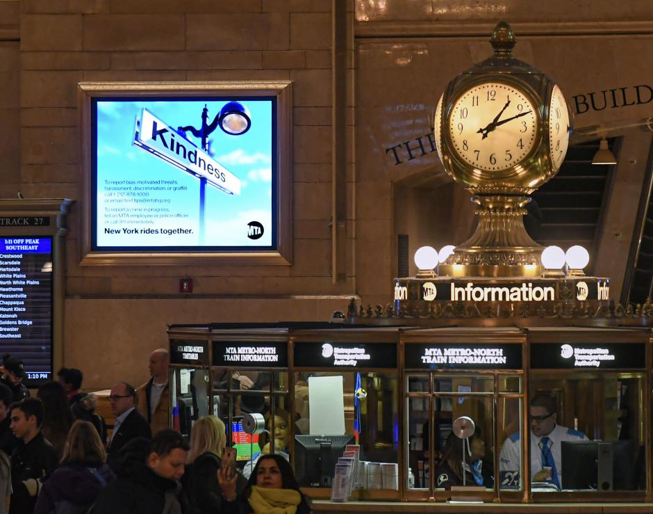 The information booth in Grand Central Station. A gold clock on the right rises above the booth. The booth has people clustered around it. On the left, a digital screen displays the word "kindness"